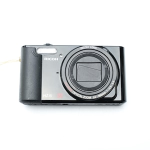 Puede incluir: Cámara digital Ricoh HZ15 negra con lente plateada y flash incorporado. La cámara tiene una correa de muñeca y está sobre un fondo blanco. La lente indica "Zoom óptico 24-360mm 1:3.3-5.9".