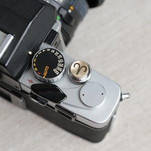 Peut inclure: Un appareil photo argenté et noir avec un cadran argenté qui indique "AUTO" et comporte des chiffres de 2 à 64. L'appareil photo a un bouton argenté avec un symbole doré du signe du zodiaque Bélier.