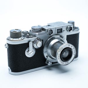 Op de afbeelding: Een vintage zilveren en zwarte camera met een prominente lens en meerdere draaiknoppen. De camerabody heeft een getextureerde zwarte grip en de lens is gemarkeerd met metingen. Een klassiek fotografie-apparaat.