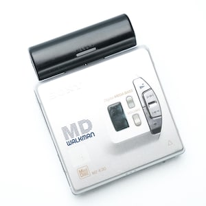 Könnte beinhalten: Silberner tragbarer digitaler Musikplayer Sony MD Walkman MZ-E30 mit schwarzem Batteriefach. Der Player verfügt über ein Bedienfeld mit Tasten für Lautstärke, Wiedergabe und andere Funktionen. Der Text "MD WALKMAN" ist sichtbar.