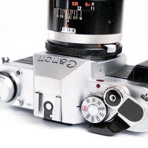 May include: Silver Canon camera with a black lens and a black leather strap. The camera has various dials and settings visible, including an ISO dial and a shutter speed dial. The word "Canon" is printed on the top of the camera.
