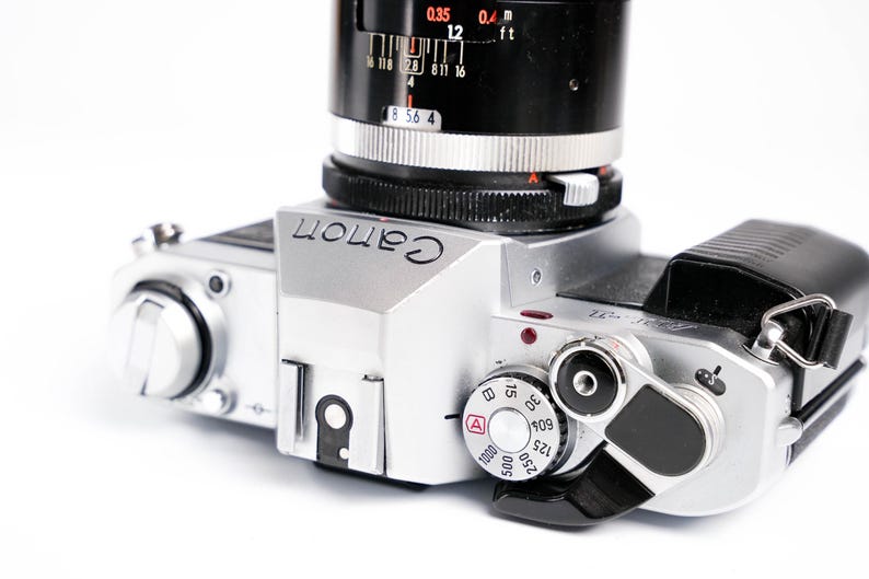 May include: Silver Canon camera with a black lens and a black leather strap. The camera has various dials and settings visible, including an ISO dial and a shutter speed dial. The word "Canon" is printed on the top of the camera.