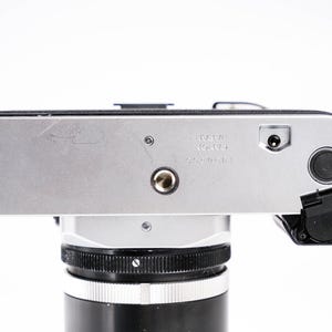 May include: Silver and black vintage camera body with a lens attached. The camera has a silver top plate with the text "MADE IN JAPAN" and a black lens. The camera is on a white background.