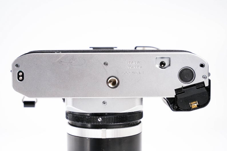May include: Silver and black vintage camera body with a lens attached. The camera has a silver top plate with the text "MADE IN JAPAN" and a black lens. The camera is on a white background.