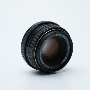 Puede incluir: Un objetivo de cámara negro SMC Pentax-M 1:2 50mm. El objetivo tiene un anillo de enfoque estriado y un anillo plateado con el texto "SMC PENTAX-M 1:2 50mm" y "ASAHI OPTICAL CO." El objetivo está ligeramente inclinado hacia la izquierda.