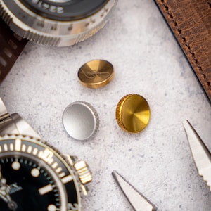 Puede incluir: Primer plano de un reloj plateado con esfera negra y bisel plateado. El reloj está rodeado de tres pequeños objetos redondos: dos dorados y uno plateado. Los objetos dorados tienen una superficie texturizada y el objeto plateado es liso.