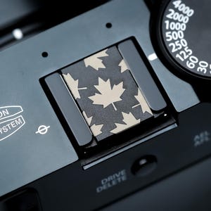 May include: Close-up of a black camera with a patterned flash shoe featuring beige maple leaves on a dark background. The camera has a dial with white numbers and the text "FUJINON LENS SYSTEM".