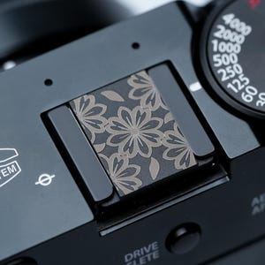 May include: Close-up of a black camera with a floral design on the top. The camera has a dial with numbers and the text "FUJINON LENS SYSTEM". The floral design is in shades of gray and brown.