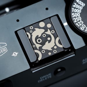May include: A close-up shot of a black camera with a silver dial and a decorative plate featuring a fish design. The camera has the text "FUJINON LENS SYSTEM" and a dial with numbers. The plate has a fish design.