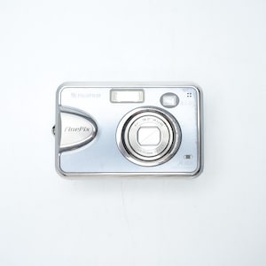 May include: A silver Fujifilm FinePix digital camera featuring a central lens, a rectangular flash unit, and a viewfinder. The front panel displays the FinePix logo on the left side and technical lens specifications printed around the glass lens element.