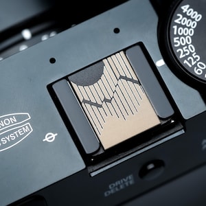 Op de afbeelding: Close-up van een zwarte camera met een zilver en beige ontwerp aan de bovenkant. De camera heeft een draaiknop met nummers en tekst, waaronder "4000" en "FUJINON LENS SYSTEM". Het ontwerp van de camera bevat een bergachtige afbeelding.