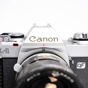 May include: Close-up of a Canon AL-1 camera. The camera body is silver and black, with the Canon logo prominently displayed. The lens is black, and the camera has various dials and buttons.