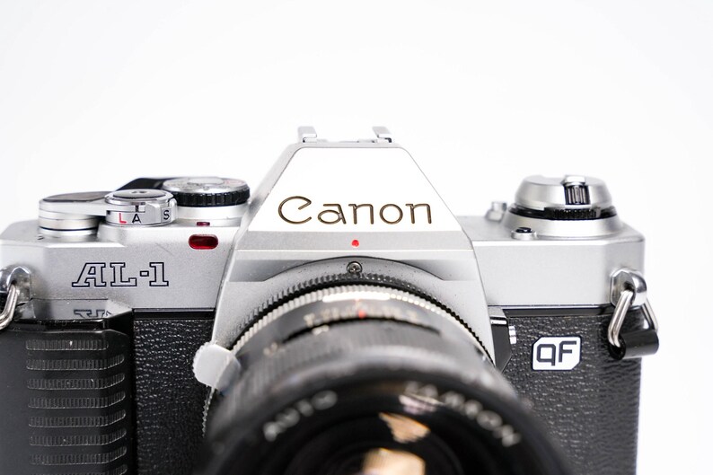 May include: Close-up of a Canon AL-1 camera. The camera body is silver and black, with the Canon logo prominently displayed. The lens is black, and the camera has various dials and buttons.