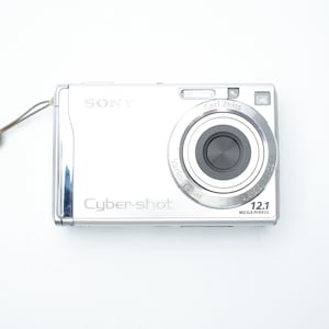 Puede incluir: Cámara digital Sony Cybershot plateada con lente Carl Zeiss y resolución de 12,1 megapíxeles. La cámara tiene una lente retráctil y una correa para la muñeca. La cámara está sobre un fondo blanco.