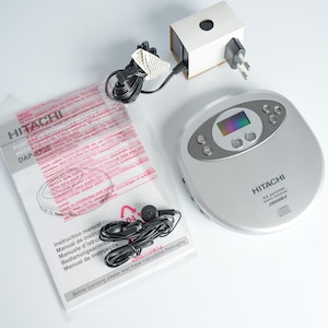 Könnte beinhalten: Ein silberner tragbarer CD-Player von Hitachi mit Digitalanzeige, Tasten und dem Text "HITACHI". Enthalten sind ein Netzteil, eine Bedienungsanleitung und Ohrhörer. Die Bedienungsanleitung trägt den Text "HITACHI DAP-XP2E".