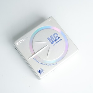 Könnte beinhalten: Ein silberner Sony MiniDisc Walkman MZ-E62 Player. Der tragbare Musikplayer hat ein kreisförmiges Design mit dem Text "MD WALKMAN Digital MEGA BASS". Der Player ist quadratisch mit einem holografischen Design.