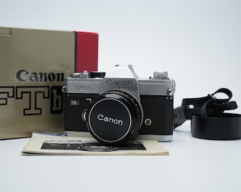 Peut inclure: Un appareil photo Canon FTb argenté avec un objectif noir et une sangle en cuir noir. L'appareil photo est posé sur une surface blanche avec une boîte brune et un morceau de papier en arrière-plan.