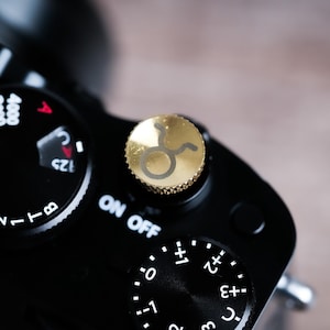 Puede incluir: Primer plano de una cámara negra con un dial dorado con el símbolo del signo zodiacal Tauro. Se ven los diales de configuración de la cámara. El fondo está borroso.
