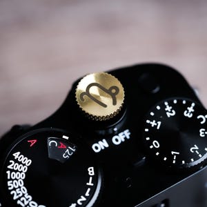 Puede incluir: Primer plano de una cámara negra con un dial dorado con un símbolo de Capricornio. La cámara tiene varios diales y ajustes, incluido un interruptor de encendido/apagado y ajustes de apertura. El fondo es una superficie de madera borrosa.