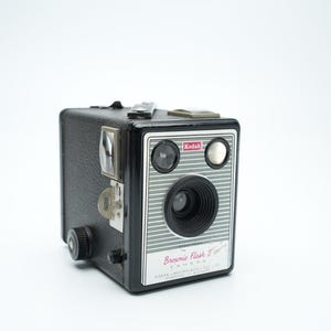 Puede incluir: Una cámara Kodak Brownie Flash II negra con un panel frontal plateado y un logotipo Kodak rojo. La cámara tiene un visor, una lente y un accesorio de flash.