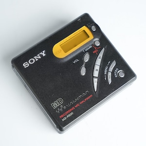 Könnte beinhalten: Schwarzer Sony MD Walkman MZ-R500. Der tragbare Audioplayer hat ein gelbes Display und silberne Tasten für Lautstärke, Menü und Markierung. Das Gerät trägt das Sony- und Walkman-Logo.