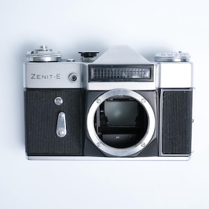 May include: A vintage Zenit-E camera, silver and black, with the model name prominently displayed. The camera features a lens mount, flash, and various dials and controls. The body has a textured grip, and the overall design is classic and functional.