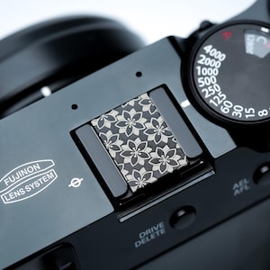 Op de afbeelding: Close-up van een zwarte Fujifilm X100VI camera. De bovenkant heeft een bloemenpatroon op de flitsschoenafdekking. De camera heeft een instelwiel voor de sluitertijd, van 8 tot 4000, en het Fujifilm Lens System logo.