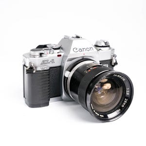 May include: Silver and black Canon AL-1 camera with a black Tamron 28mm f/2.8 lens. The camera body has the Canon logo and AL-1 model name. The lens has white text indicating its specifications.