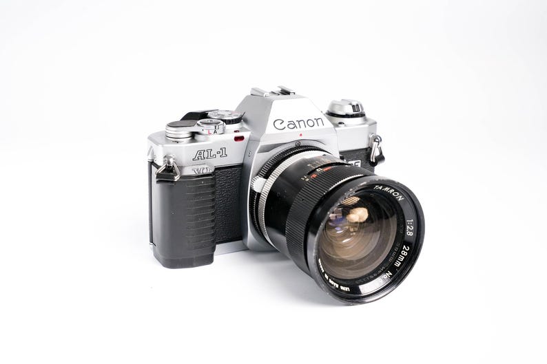 May include: Silver and black Canon AL-1 camera with a black Tamron 28mm f/2.8 lens. The camera body has the Canon logo and AL-1 model name. The lens has white text indicating its specifications.