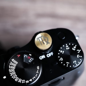May include: Close-up of a black camera with gold accents. The camera features a gold dial with the Virgo symbol and various control dials with white markings. The camera's settings include aperture, shutter speed, and power.