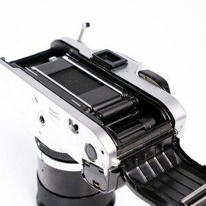 May include: A silver and black Canon camera with the back open, revealing the film compartment. The camera lens is black, and the body is silver. The open back shows the internal components, including the film rollers and the shutter mechanism.