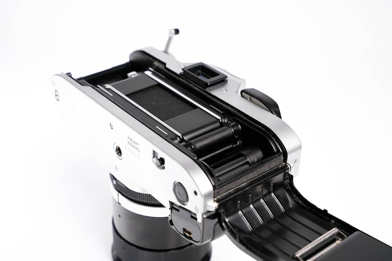 May include: A silver and black Canon camera with the back open, revealing the film compartment. The camera lens is black, and the body is silver. The open back shows the internal components, including the film rollers and the shutter mechanism.