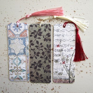 May include: Three decorative bookmarks with tassels. The bookmarks feature different designs: a tile pattern, a leafy pattern, and a floral and script design. Each bookmark has a tassel in a different color: pink, cream, and red.