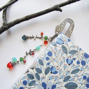 Könnte beinhalten: Ein silbernes Lesezeichen mit einem verzierten Design und einem baumelnden Anhänger mit einem floralen Motiv und bunten Perlen. Das Lesezeichen liegt auf einem Buch mit einem blauen und weißen Blumenmuster.