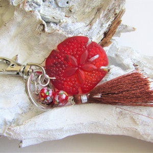 May include: Red glass sand dollar keychain with a brown tassel and a silver charm. The keychain has a silver ring with a small, colourful glass bead.
