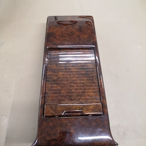 Puede incluir: Una moldura de madera para el interior de un coche con un acabado de madera de color marrón oscuro y una abertura rectangular con una tapa deslizante.