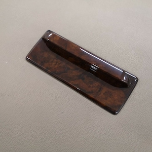 May include: A rectangular wooden trim piece with a raised, smooth, dark brown handle. The trim piece has a wood grain pattern and a glossy finish.