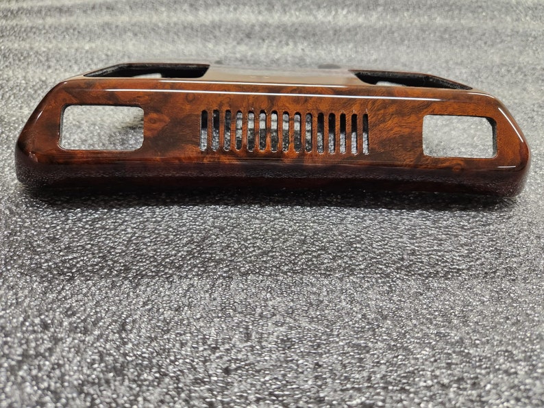 May include: A dark brown, wood-grain effect car interior trim. The trim has a glossy finish and a rectangular shape. It features two rectangular openings and a series of vertical vents in the centre.