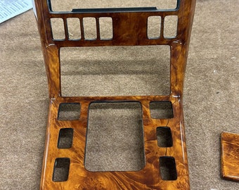 May include: A wooden car dashboard trim piece with a woodgrain pattern and multiple rectangular cutouts. The trim piece is brown with a glossy finish.