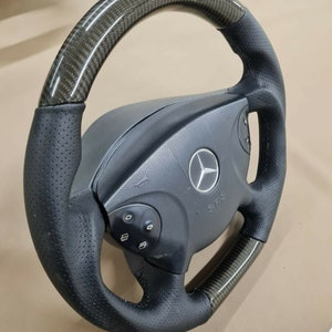 Op de afbeelding: Een zwart lederen stuurwiel met een carbonfiber afwerking. Het stuurwiel heeft een Mercedes-Benz logo en de tekst SRS.