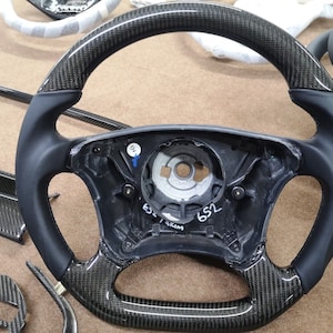 Puede incluir: Un volante de cuero negro con un borde de fibra de carbono. El volante tiene un cubo central plateado con el número "652" impreso en él.