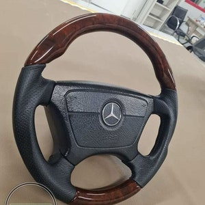 May include: A black steering wheel with a wood grain top and bottom. The center of the wheel has a silver emblem and the words "SRS AIRBAG". The wheel is likely for a car.