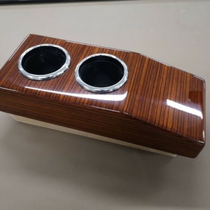 Puede incluir: Un portavasos de madera con dos portavasos con borde cromado. El portavasos tiene un acabado de veta de madera y un acabado brillante.