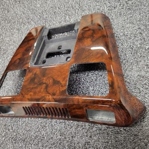 May include: A car dashboard trim with a glossy wood-grain effect. The trim is brown with darker, swirling patterns. It has cutouts for vents and controls. The shape is rectangular. This interior car accessory is designed to enhance the vehicle's aesthetics.
