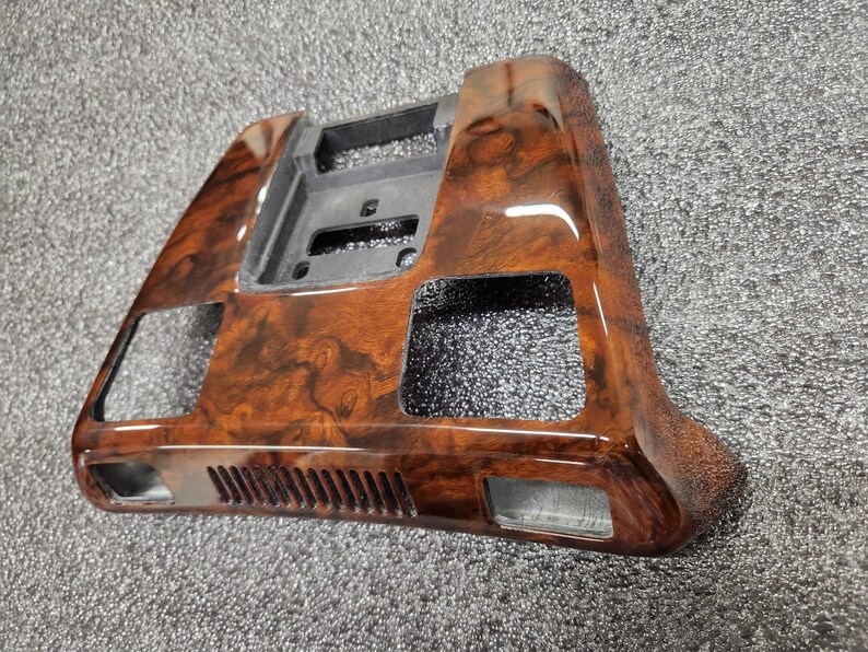 May include: A car dashboard trim with a glossy wood-grain effect. The trim is brown with darker, swirling patterns. It has cutouts for vents and controls. The shape is rectangular. This interior car accessory is designed to enhance the vehicle's aesthetics.