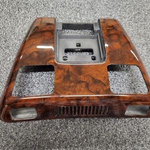 May include: A car interior trim piece with a glossy wood-grain effect. The trim is predominantly brown with darker, swirling patterns. It has rectangular openings and a central, recessed section with a black plastic component.