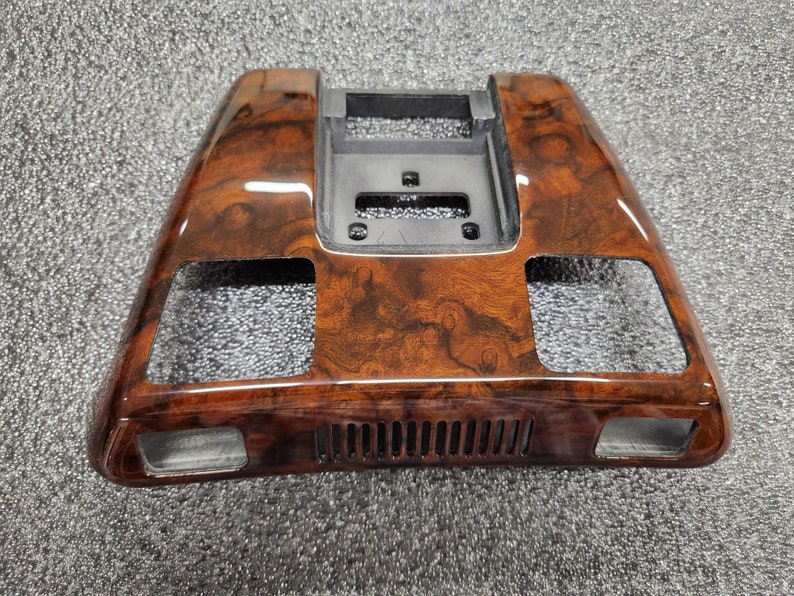 May include: A car interior trim piece with a glossy wood-grain effect. The trim is predominantly brown with darker, swirling patterns. It has rectangular openings and a central, recessed section with a black plastic component.