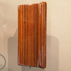 Puede incluir: Panel rectangular de madera con acabado brillante. El panel presenta un patrón a rayas en tonos marrones y rojizos. Los bordes son redondeados y parece ser parte de una estructura o mueble más grande.