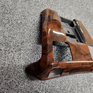 May include: A car interior trim piece with a glossy, wood-grain finish. The trim is brown with darker, swirling patterns. It has a rectangular shape with cut-out sections and a black plastic backing.