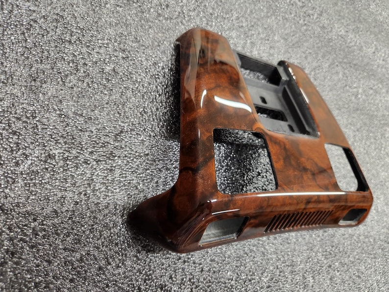 May include: A car interior trim piece with a glossy, wood-grain finish. The trim is brown with darker, swirling patterns. It has a rectangular shape with cut-out sections and a black plastic backing.
